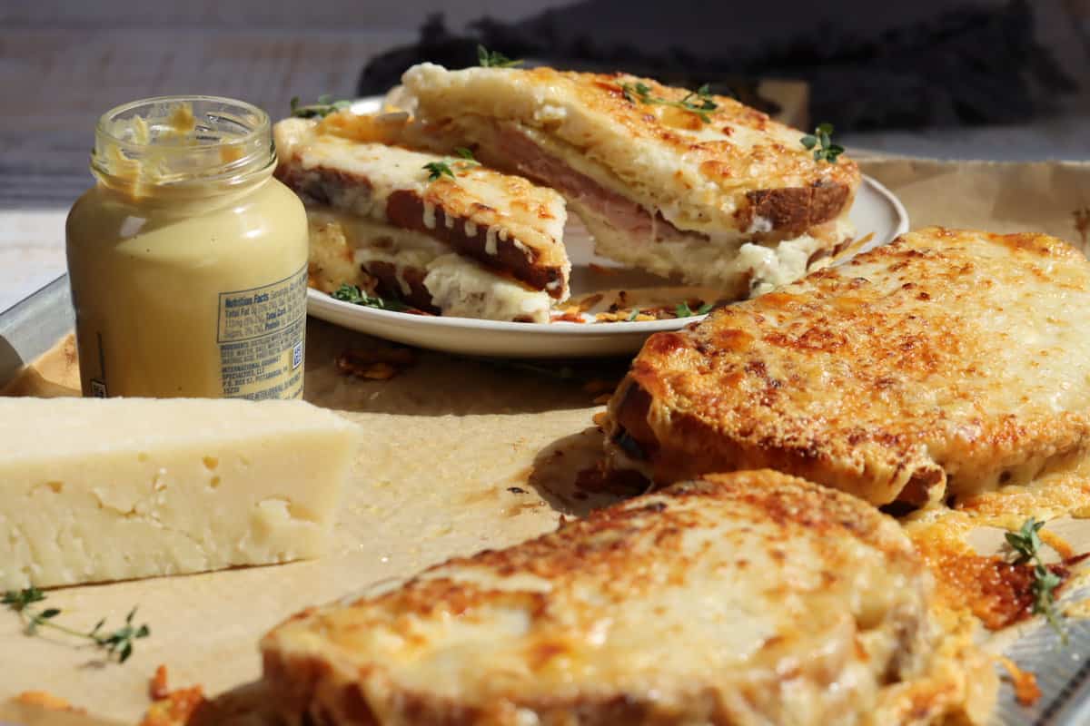 cheesy crispy croque monsieur sandwiches on a sheet pan and one cut in half on a plate