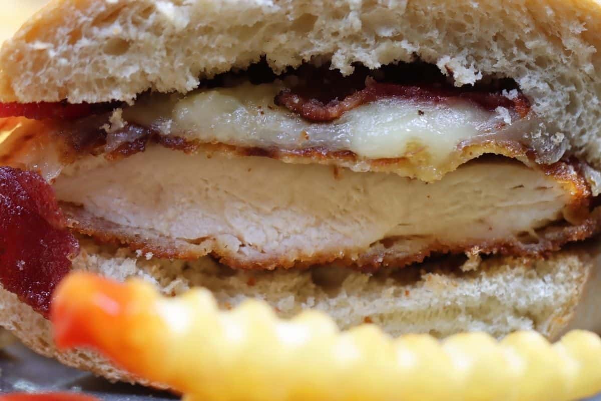 extreme close up of chicken cutlet with bacon and cheese cut in half on french bread plus a french fry dipped in ketchup