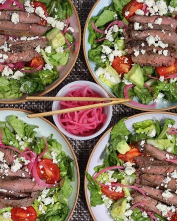 overhead shot of four steak salads with pickled red onions and bleu cheese as well as campari tomatoes, avocado chunks and homemade balsamic vinaigrette
