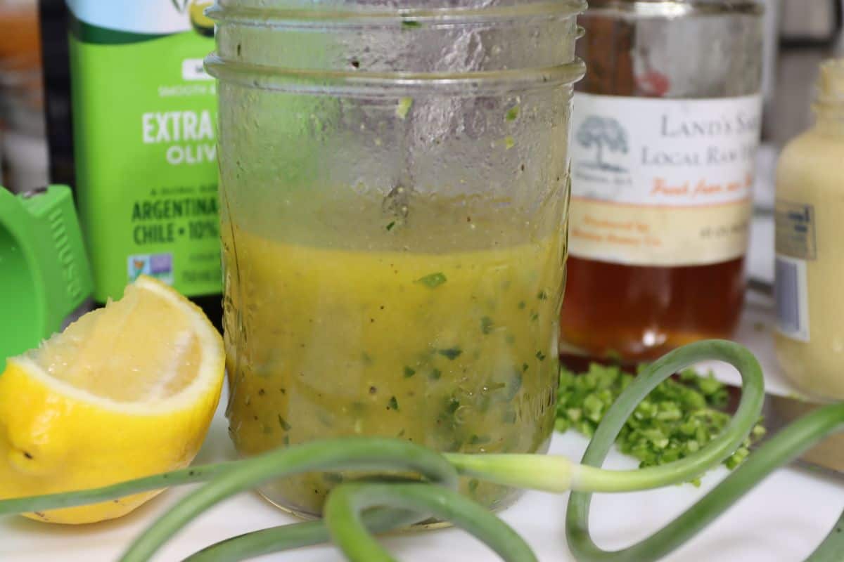 mason jar filled with super tangy garlic scape vinaigrette with garlic scapes, lemon, olive oil, mustard and honey in background on cutting board