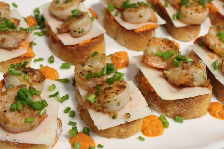 side view of garlic shrimp crostini with romesco sauce and shaved parmesan topped with fresh chives and garnished with dots of sauce