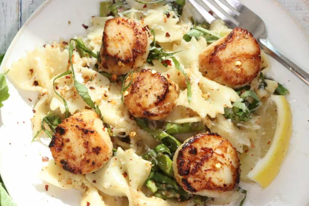 one plate of pan seared sea scallops with basil brown butter, asparagus and farfalle pasta garnished with fresh basil, parm and red pepper flakes