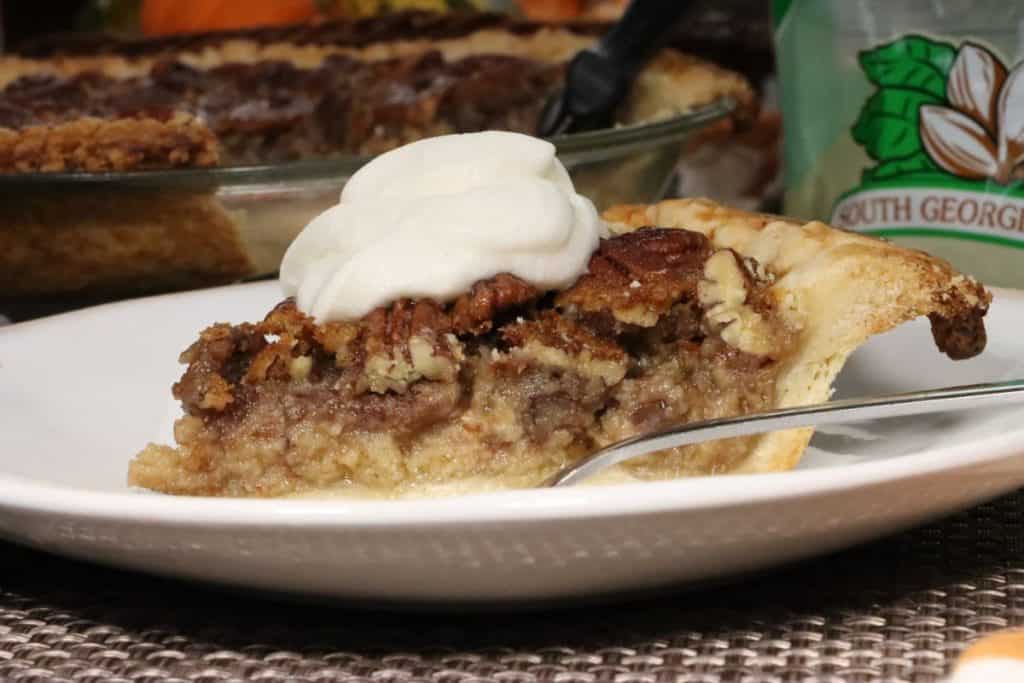 just a little slice of heaven on a plate with this bourbon pecan pie topped with whipped cream. The whole pie is in the background and a bag of pecan on the table