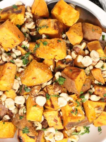 overhead view of a serving bowl filled with roasted sweet potatoes with toasted hazelnuts, a real maple syrup drizzle and fresh parsley