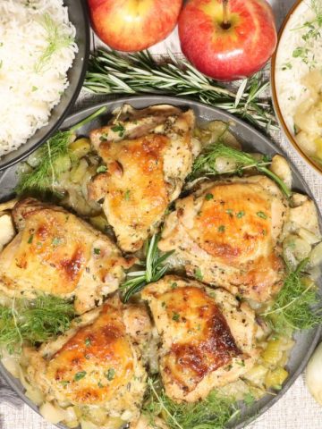 Dijon Chicken with Apples, Leeks and Fennel: overhead view of platter of chicken on the table with one portion on a plate to the side, bowl of basmati rice, apples, rosemary, fennel on the table
