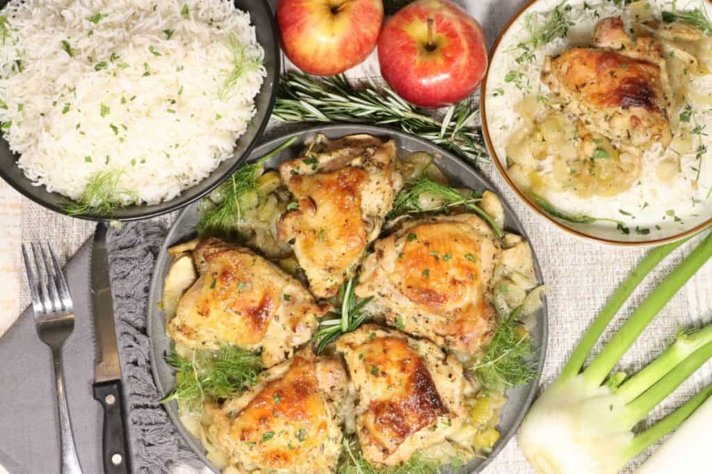 Dijon Chicken with Apples, Leeks and Fennel: overhead view of platter of chicken on the table with one portion on a plate to the side, bowl of basmati rice, apples, rosemary, fennel on the table