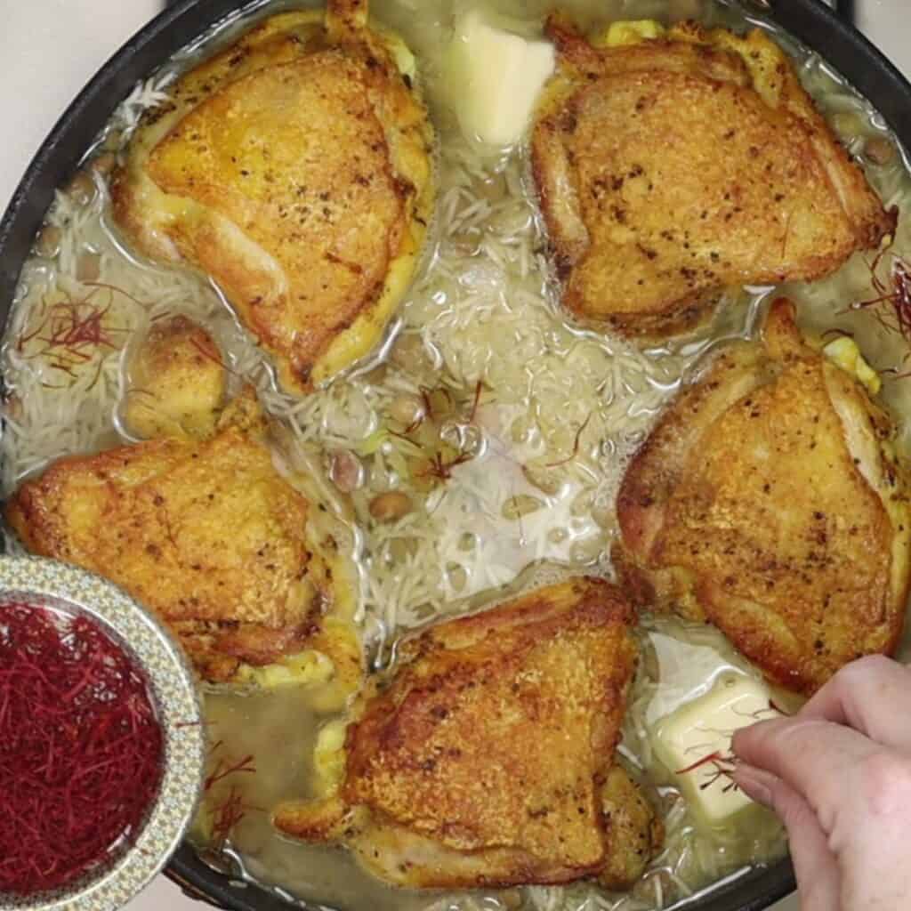 two pats of butter and inches of saffron being added to crispy saffron chicken thighs with barberries and pigeon peas