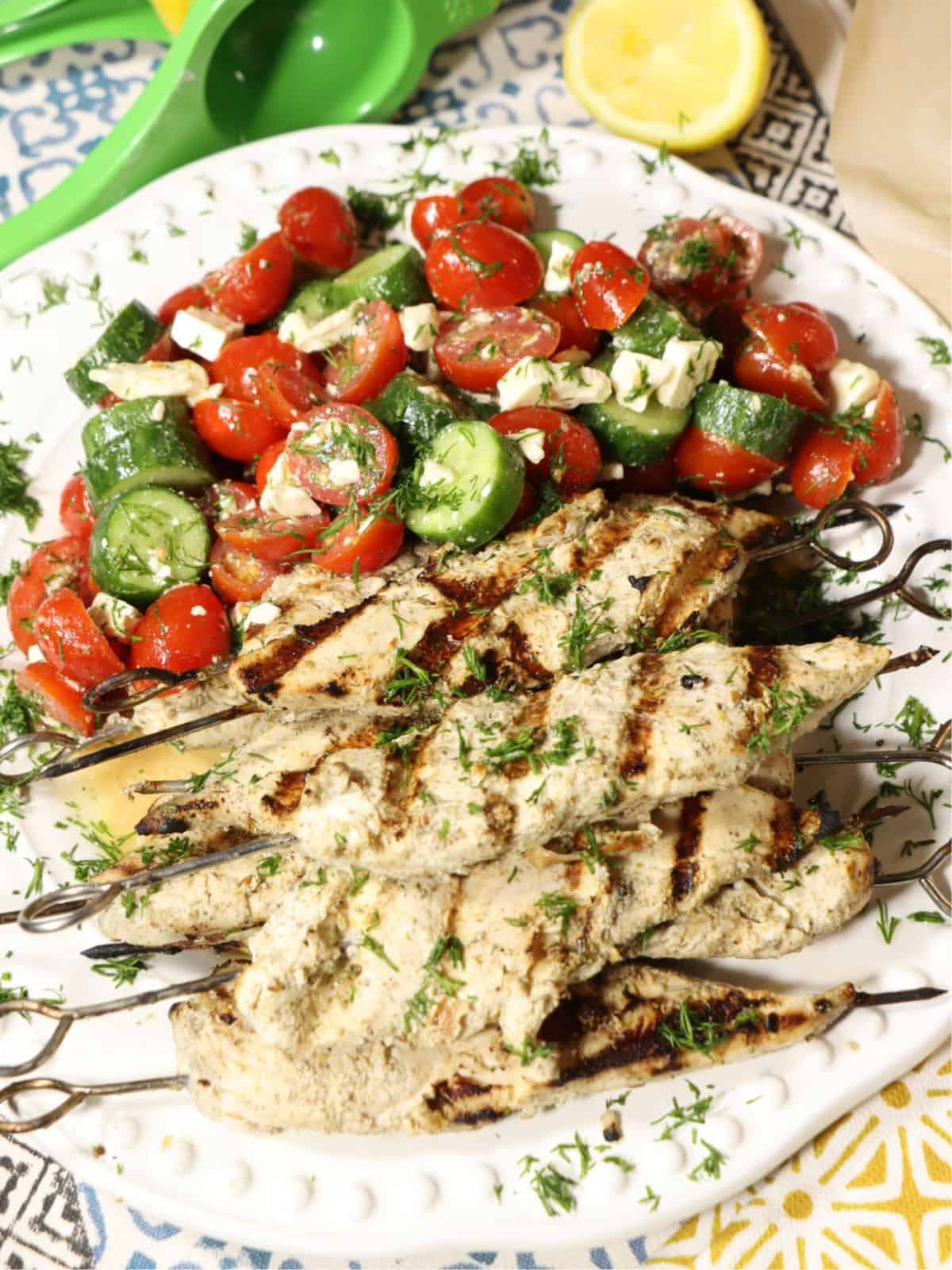 side view of grilled za'atar lemon chicken skewers on a plate with tomato, cucumber, dill salad