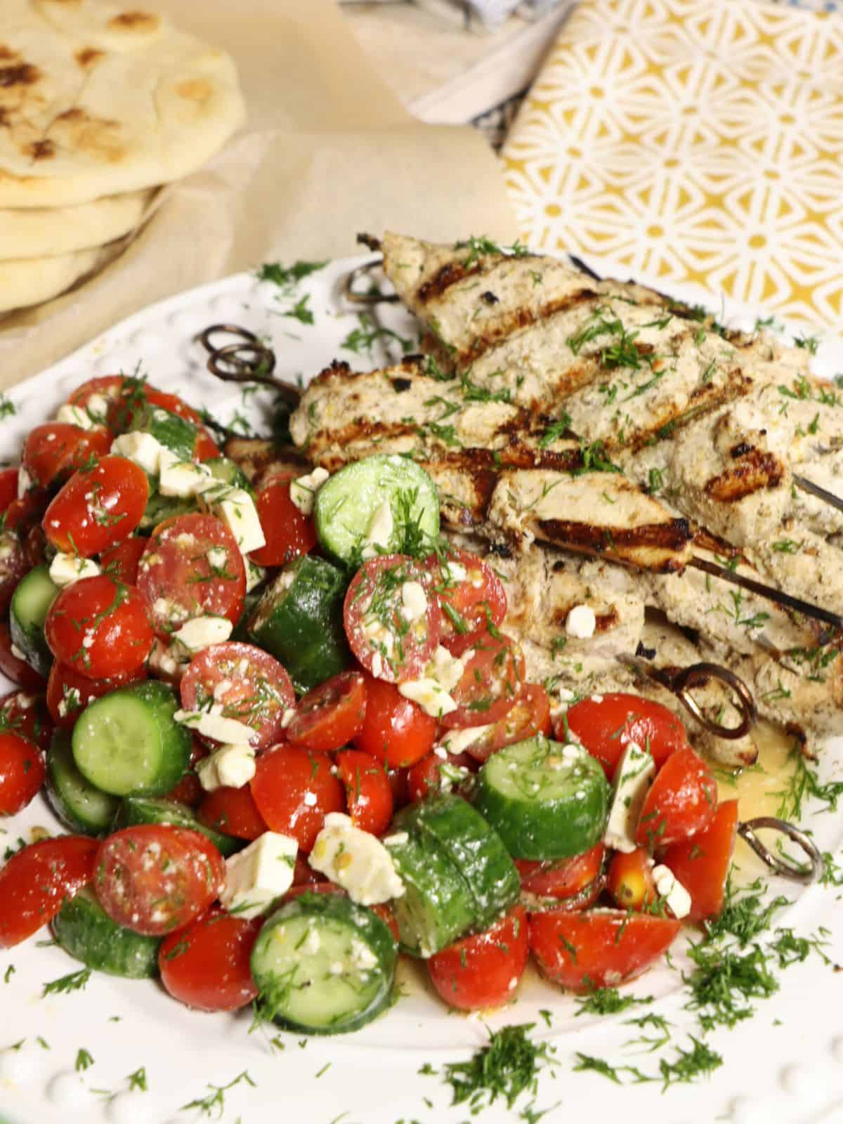 close up front view of grilled za'atar lemon chicken skewers on a plate with tomato, cucumber, salad